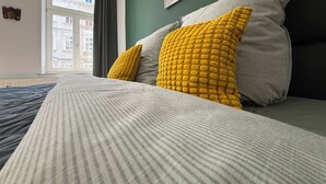 Un letto con lenzuola a righe e due cuscini, uno giallo e uno grigio, in una stanza con una parete verde e una finestra.