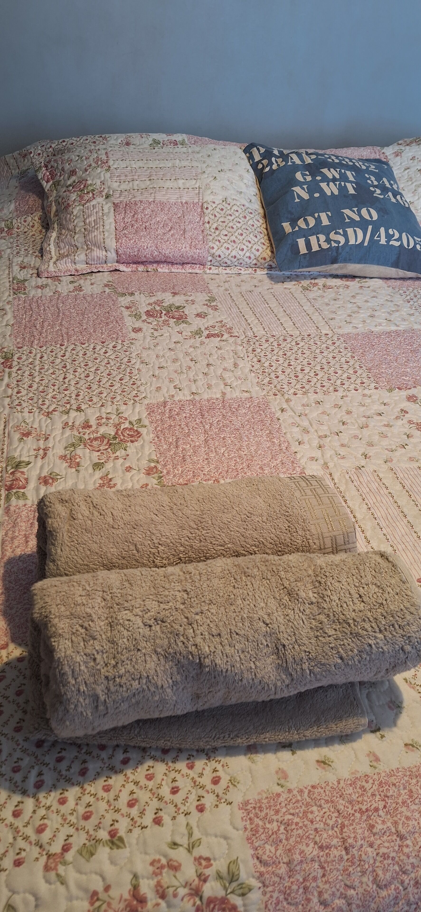 Una cama con colcha floral, un almohón azul con texto y una manta doblada.