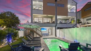Un edificio moderno de dos pisos con terraza en la azotea, zona de estar exterior con sillones y piscina.