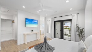 Una habitación de hotel moderna con una cama grande, un ventilador de techo, vistas al exterior, una mesita con una planta y un cuadro en la pared.