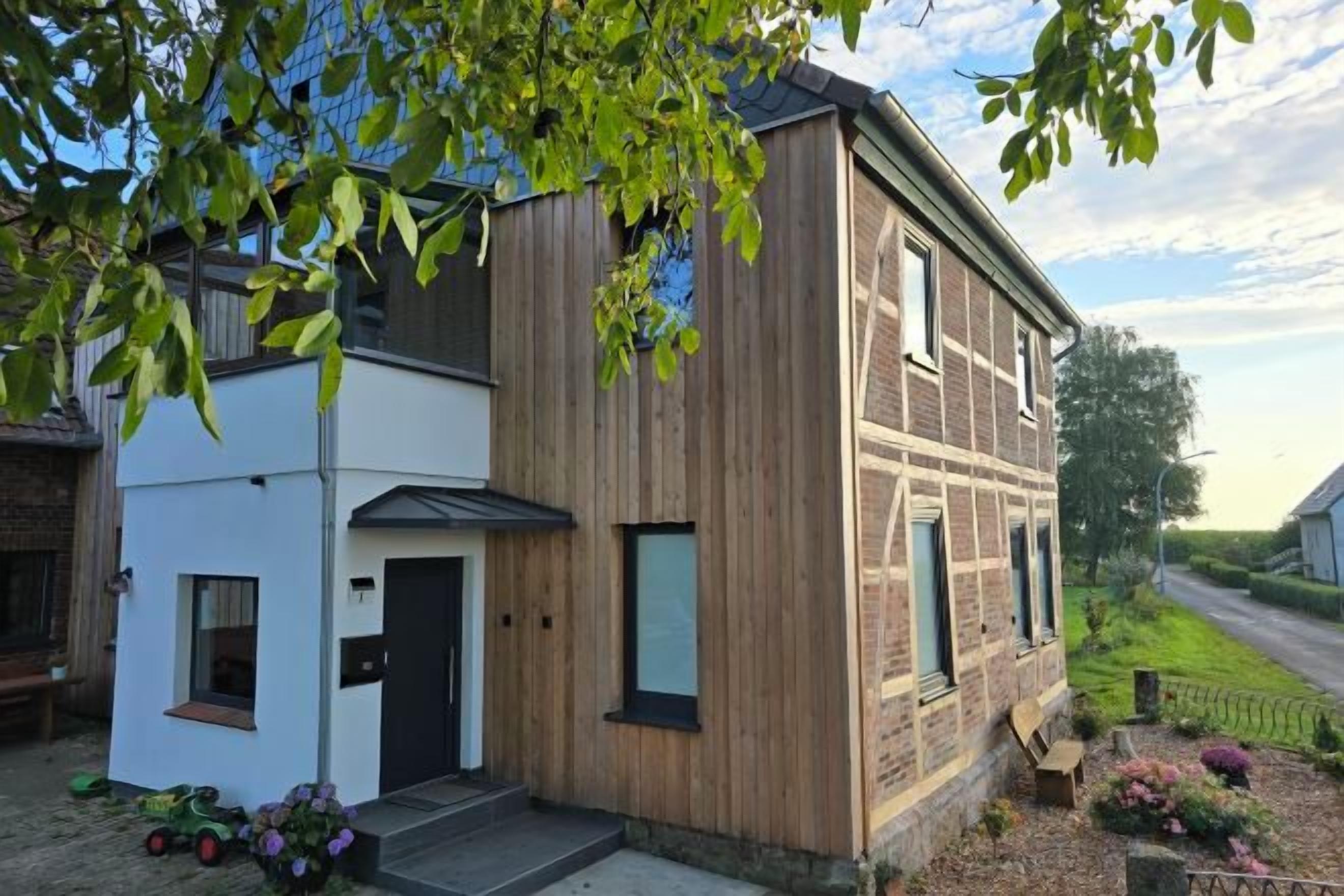 A two-story building with a wooden facade, a small attached white structure, and a paved entrance area with flowers.