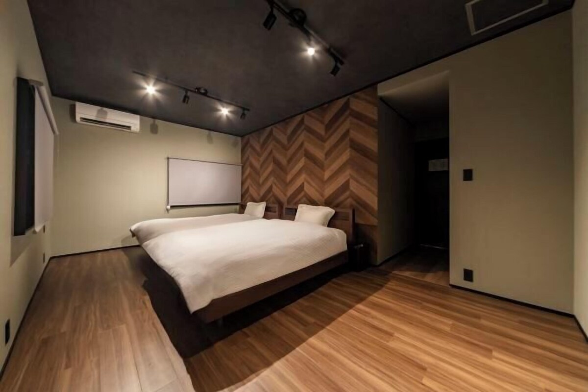A modern bedroom with a large bed, wooden flooring, a herringbone-patterned wall, and recessed ceiling lighting.