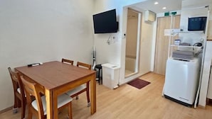 A compact living space with a wooden dining table, a flat-screen TV mounted on the wall, a white refrigerator, and a small kitchenette area. - Shikai Family (101) (Osaka)
