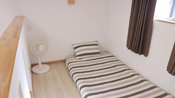 A small, single-bed room with a striped bedspread, a standing fan, and a window with curtains. - Shikai Family (101) (Osaka)