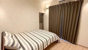 A single bed with a striped bedspread, a wooden headboard, a small bedside table, a wall-mounted air conditioner, and a window with curtains. - Shikai Family (101) (Osaka)