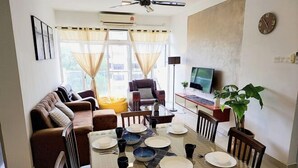 A modern living room with a sofa set, dining table, and chairs. There is a television on a stand, a plant, and a clock on the wall. - Putrajaya Dwiputra 6 Plus 1 PAX Fully Air-Con (Putrajaya)