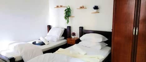A hotel room with two single beds, a wardrobe, a bedside table with a lamp, and a small shelf with decorative items.