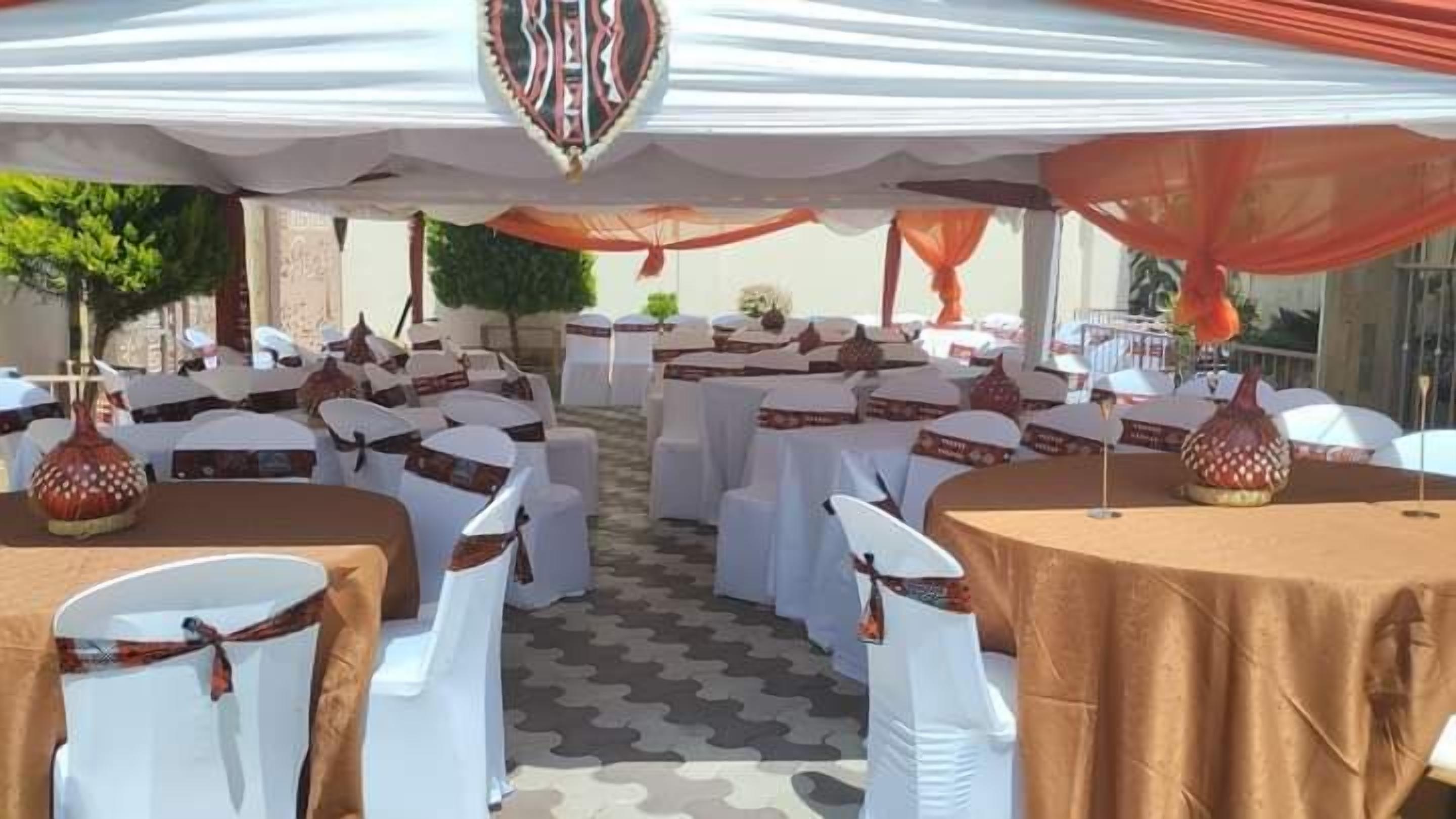 An outdoor event space with round tables covered in white tablecloths and white chairs with brown sashes.