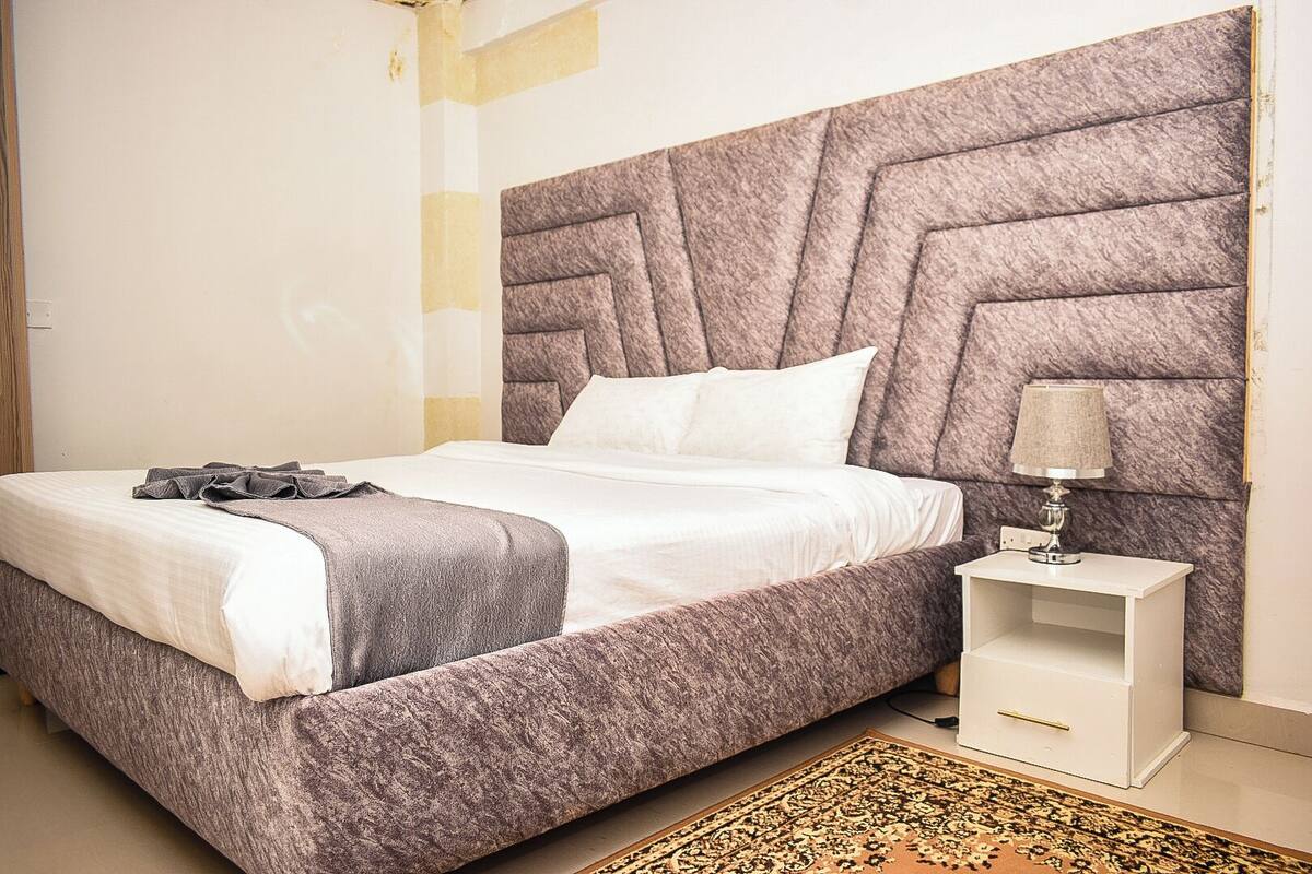 Een moderne slaapkamer met een groot bed, een hoofdbord met een geometrisch patroon, een nachtkastje met een lamp en een tapijt met dessin.