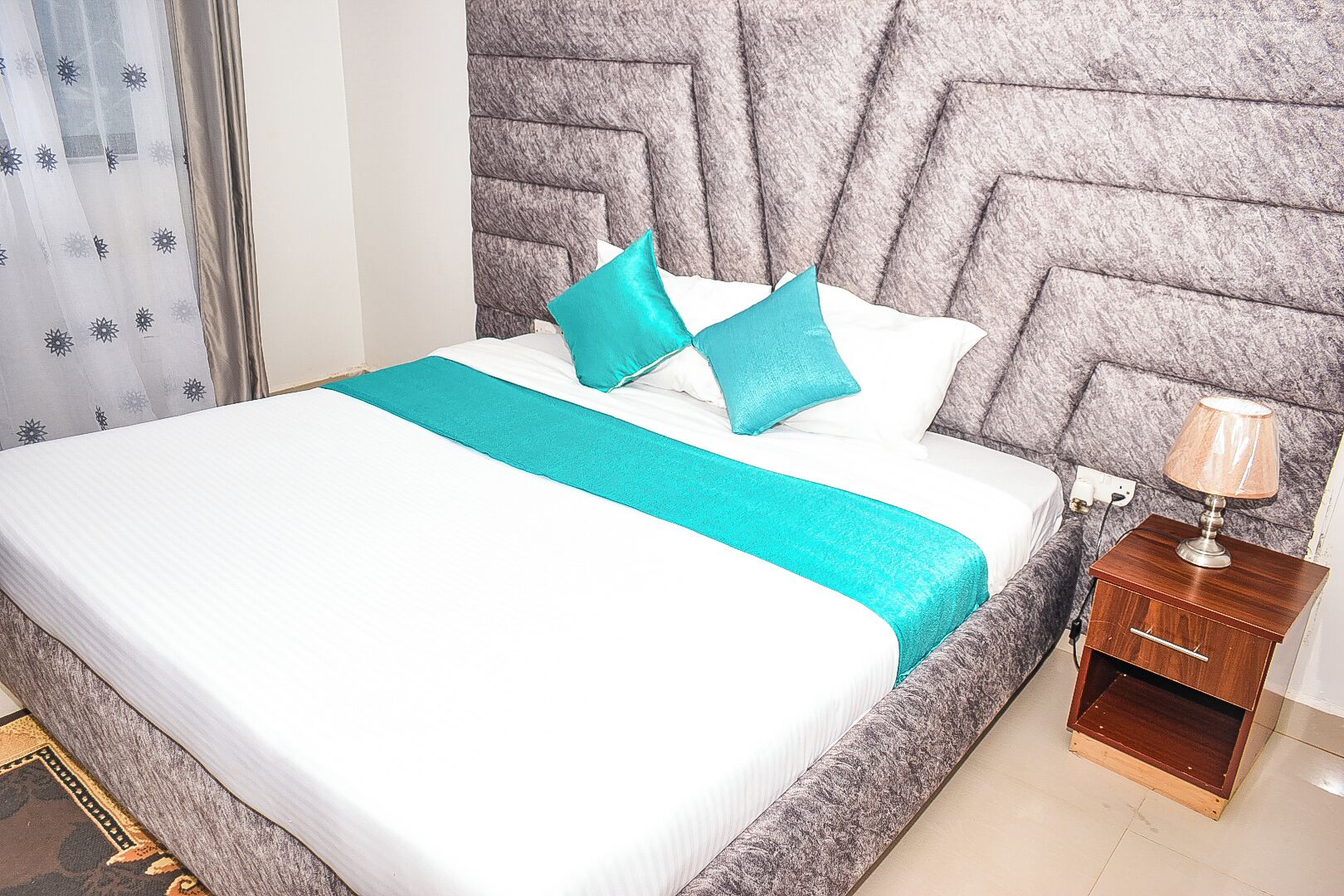 A neatly made bed with a white and teal bedspread, a grey headboard, and a wooden nightstand with a lamp.