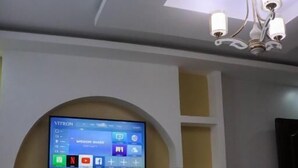 Un salon avec un téléviseur fixé au mur qui affiche différentes applications, un luminaire au plafond et une petite table sur laquelle se trouve une bouilloire.