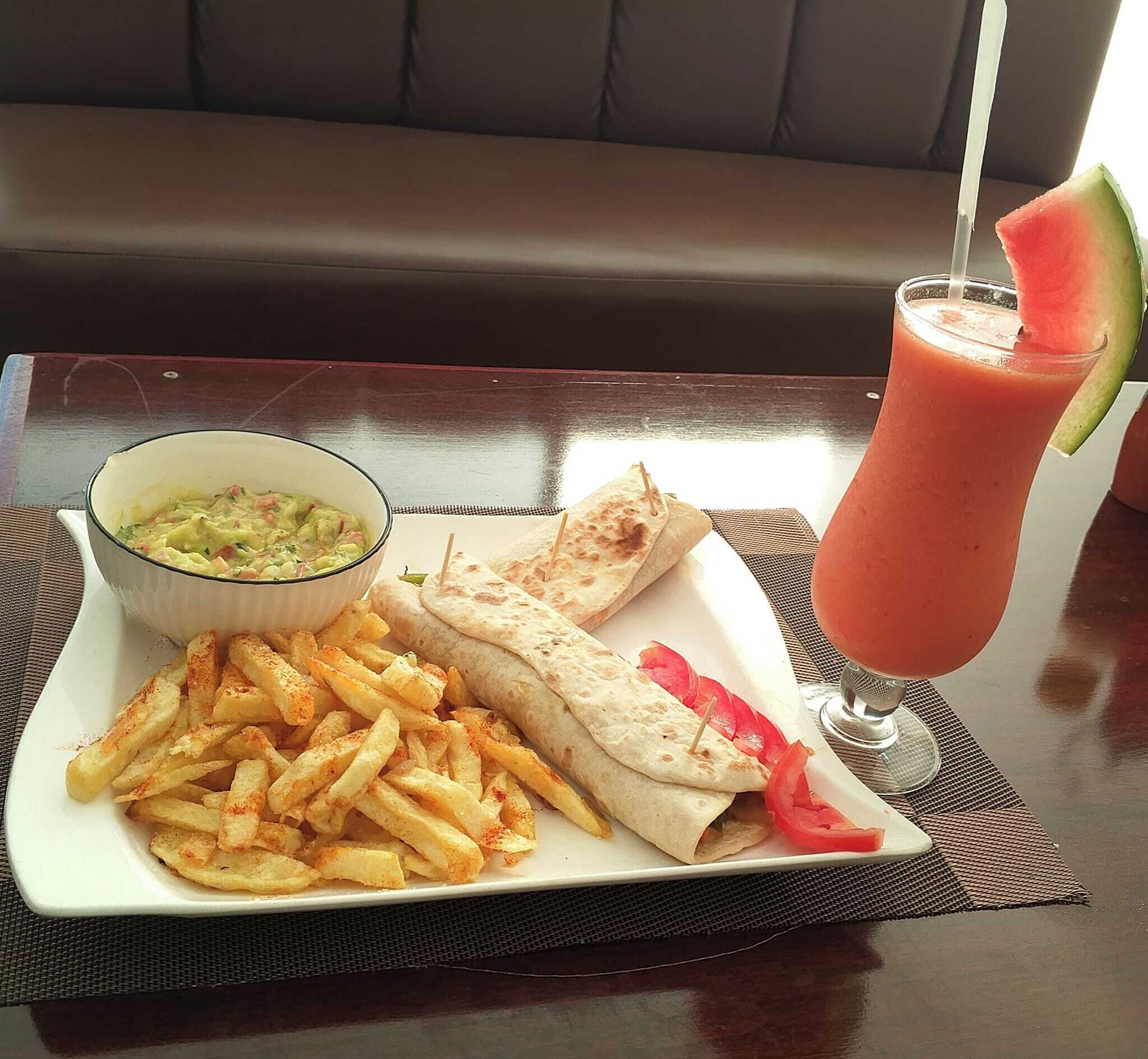 Un plato con papas fritas, un wrap, un tazón de sopa y un vaso de batido con una tajada de sandía.