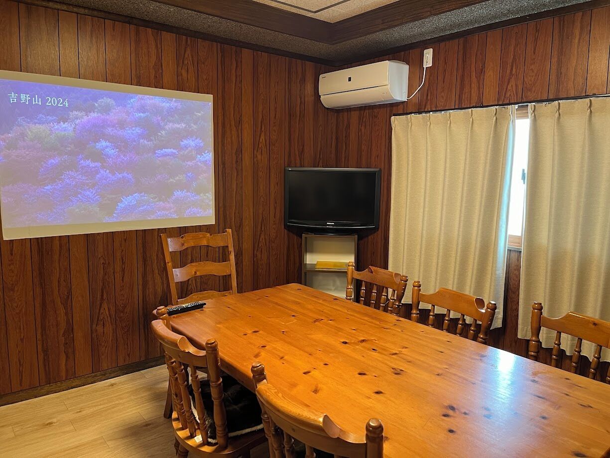 Un salón de conferencias con una mesa de madera, sillas, una pantalla de proyector, un televisor de pantalla plana y un equipo de aire acondicionado.
