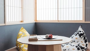 Une pièce avec une table ronde, une fenêtre avec des stores et deux coussins à motifs.