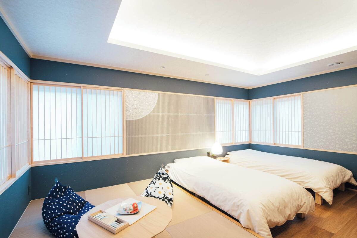 Une pièce avec un sol recouvert de tatamis, un lit avec une literie blanche, une petite table avec une lampe et des paravents en papier de riz (shoji).