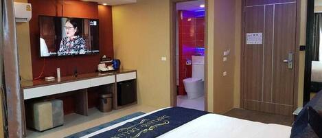 A hotel room with a bed, a TV mounted on the wall, a small desk with a chair, and a door leading to a bathroom.