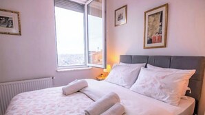 Ein ordentlich gemachtes Bett mit weißen Leinen und aufgerollten Handtüchern, eine Nachttischlampe und ein Fenster mit Blick auf Gebäude.