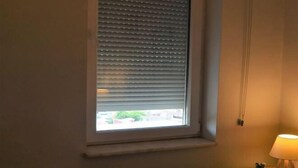 A room with a window with horizontal blinds, a bedside table with a lamp, and a small plant. - Gorgeous Two Bedroom Apartment - Roya (Istanbul)