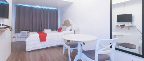 Quarto de hotel com cama, mesa redonda, duas cadeiras, televisão e espelho grande.