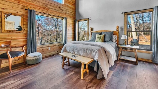 Une chambre située dans une cabane en bois, avec un lit, un banc, une chaise et de grandes fenêtres donnant sur la forêt.