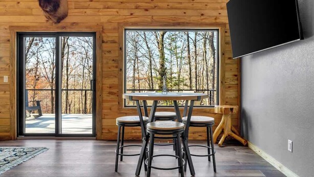 Un interior de madera con una mesa redonda y cuatro taburetes, ventanas amplias con vistas a un bosque y un televisor de pantalla plana montado en la pared.