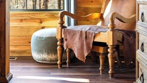 Interior de una cabaña de madera con una silla de madera, un reposapiés acolchado y un aparador.