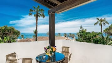 Ein überdachter Sitzbereich im Freien mit Tisch und Stühlen, Blick auf das Meer und Palmen.