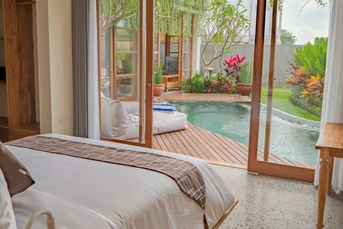 Ein Schlafzimmer mit Bett, Holzboden, Glastür zu einem Poolbereich mit Pflanzen und einem Fernseher.
