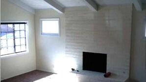 Une pièce avec une cheminée en briques blanches, une fenêtre avec des barreaux et un plancher recouvert d’un simple tapis.