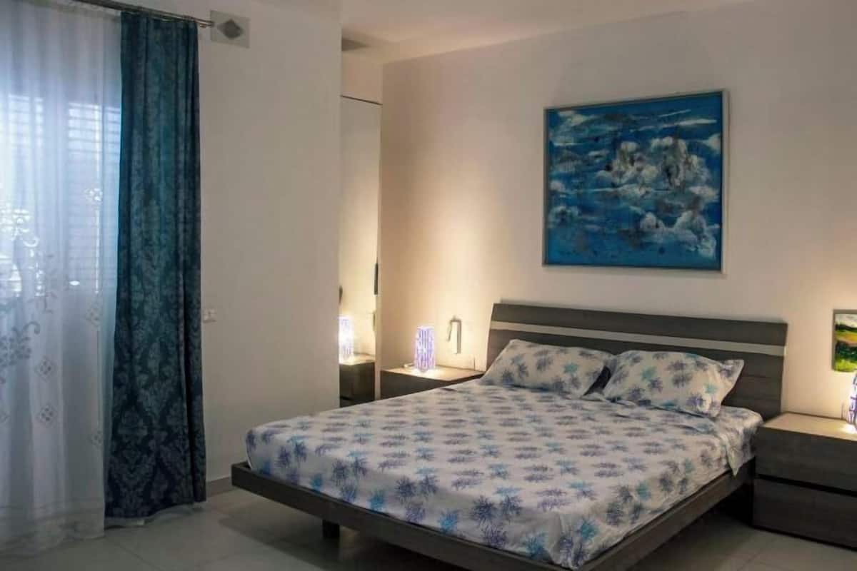 A bedroom with a bed, a nightstand, a wall-mounted lamp, and a painting.