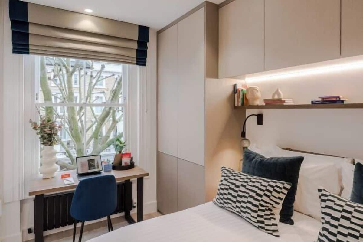 Une chambre moderne avec un lit, une table de chevet, une lampe et une fenêtre donnant sur des immeubles.
