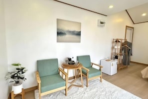 Una habitación con dos sillas verdes, una mesita redonda pequeña, un radiador blanco, una planta en maceta y un cuadro en la pared.