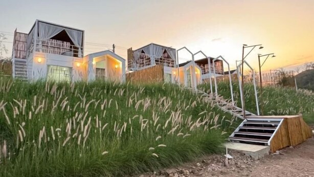 Een reeks moderne, witte, meerlaagse cabins met balkons, tegen een achtergrond van hoog gras en een zonsondergang.