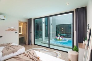 A modern hotel room with a large sliding glass door leading to a pool area, a bed with a blanket, a bedside table, and a chair. - Nacha Pool Villa Cha-am (Cha-am)