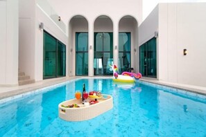 An indoor swimming pool with clear glass walls, a floating bar area with drinks, and a colorful inflatable pool float. - Nacha Pool Villa Cha-am (Cha-am)