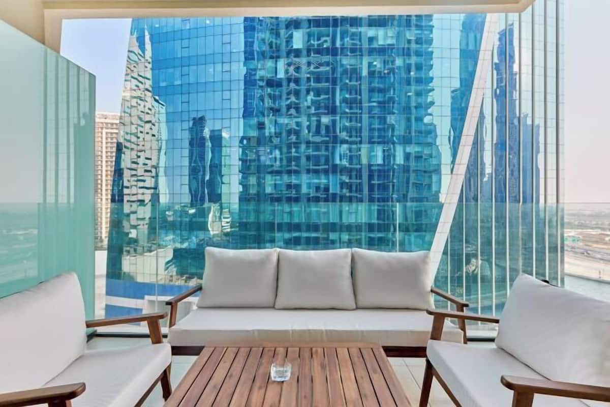 Un balcón con sofá y dos sillones, con vista a un paisaje urbano de edificios modernos.