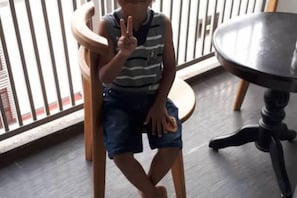 A child sitting on a wooden chair with a peace sign, a round table with a black top, and a window with vertical blinds. - Mahasa Homestay Kuala Lumpur (Kuala Lumpur)