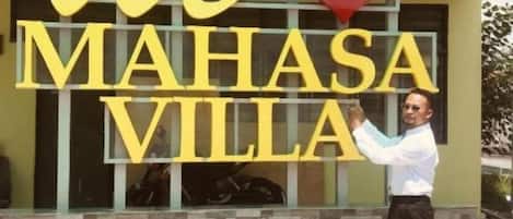 Una persona está de pie frente a un edificio con letras amarillas grandes que deletrean 'We Mahasa Villa'.