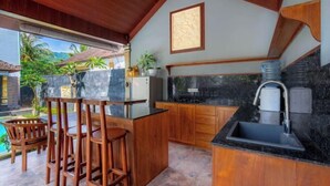 A kitchen with wooden cabinets, a black countertop, and a sink. There are two wooden stools and a small table. - Sekarini Place Amed (Bali)