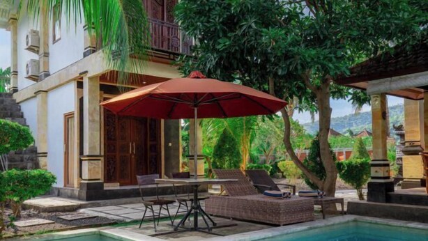 A pool area with lounge chairs and a table under a red umbrella, surrounded by lush greenery and a building with a balcony. - Sekarini Place Amed (Bali)