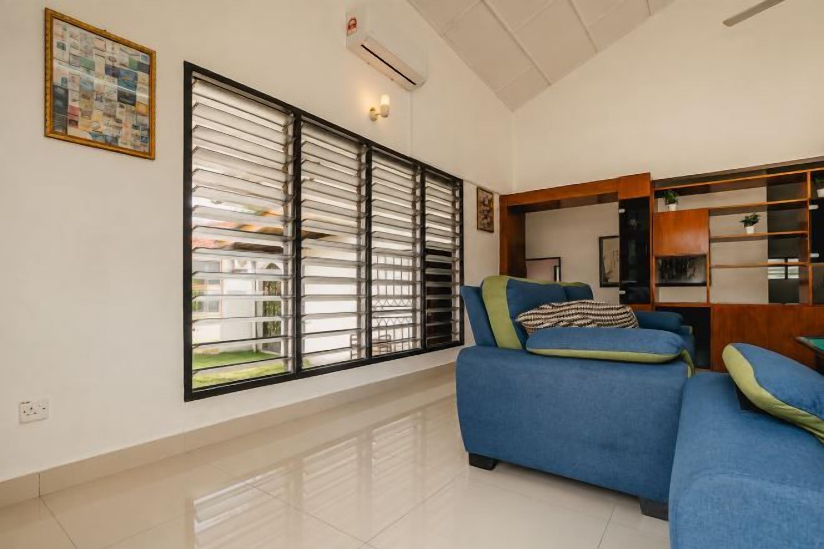 A living room with a blue sofa, a large window with blinds, and a wall-mounted air conditioner.