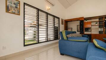 A living room with a blue sofa, a large window with blinds, and a wall-mounted air conditioner.