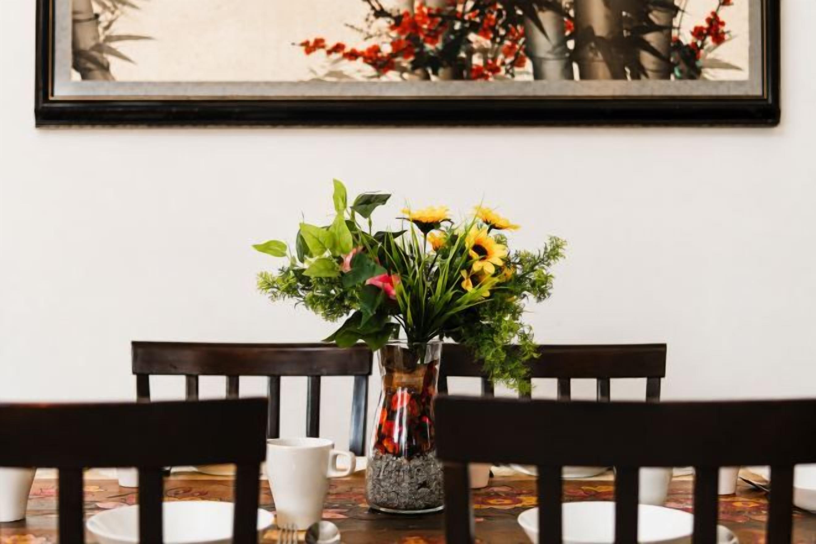 Una mesa de comedor con sillas, un florero con flores y un cuadro en la pared.