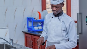 A chef in a white uniform preparing vegetables in a kitchen with a marble countertop, a sink, and a blue dish rack in the background.