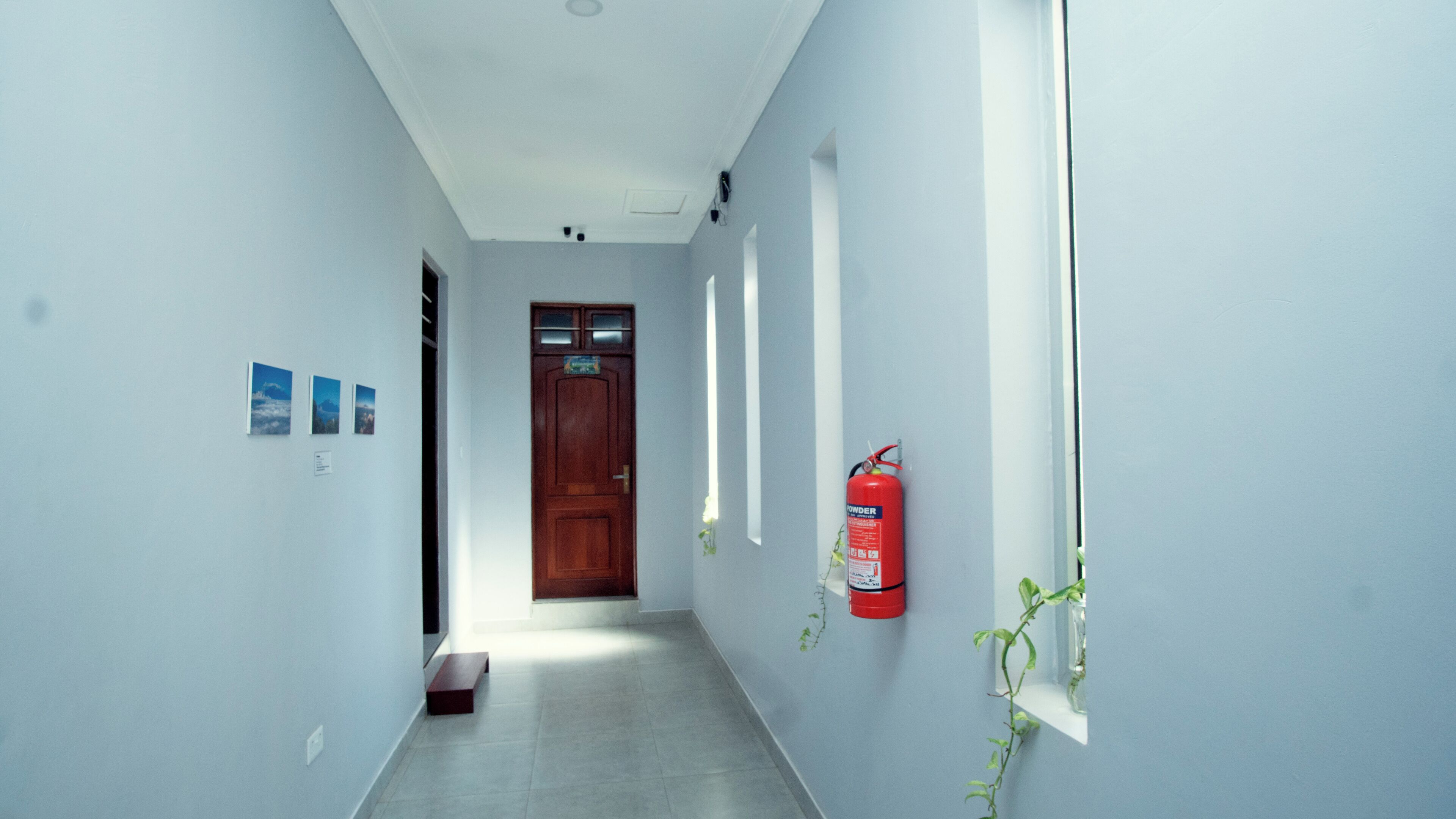 Un couloir étroit aux murs blancs, une porte en bois au fond, et un extincteur fixé au mur.