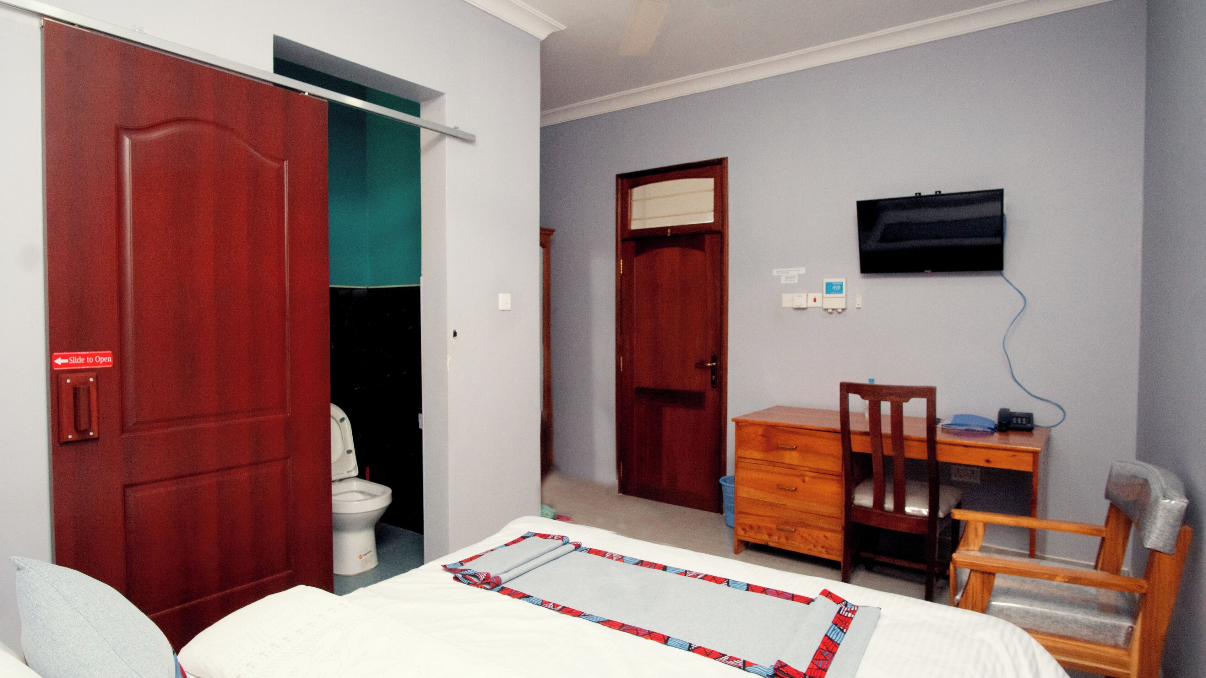 A bedroom with a bed, a desk, a chair, a television, and two doors leading to a bathroom and another room.