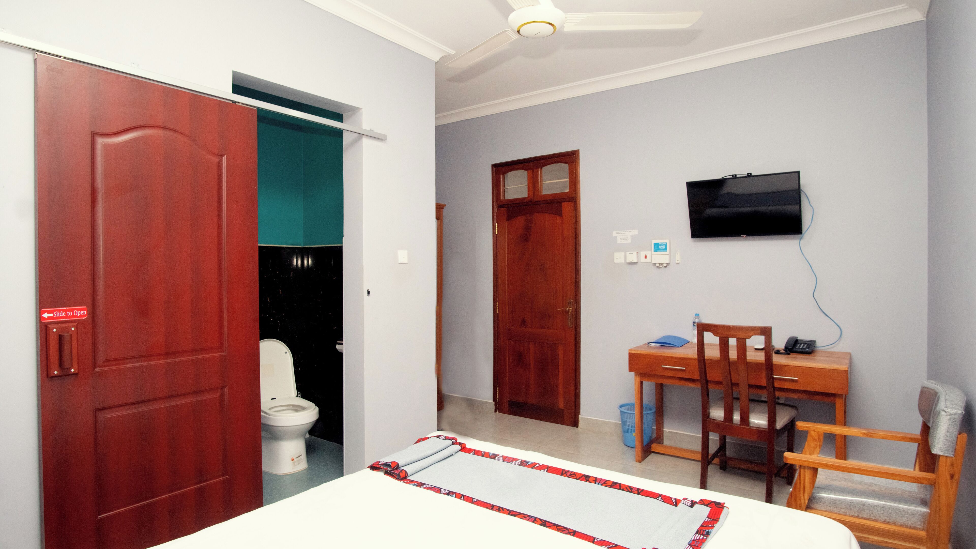 A bedroom with a bed, a desk with a chair, a TV, and a toilet.