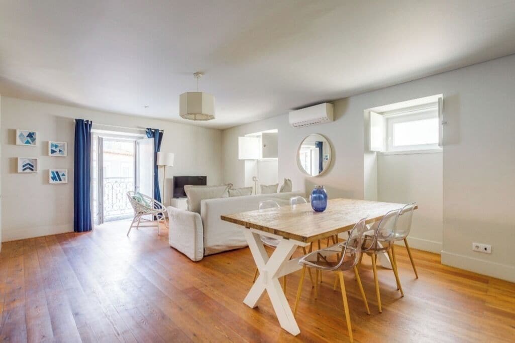 Un amplio salón con piso de madera, un sofá blanco, una mesa de comedor con sillas y un ventanal con cortinas.