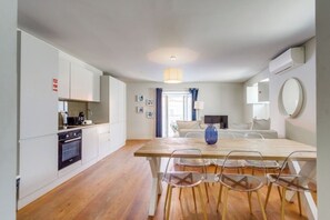 Cozinha moderna com armários brancos, uma mesa de jantar de madeira com cadeiras transparentes e uma janela grande com cortinas.