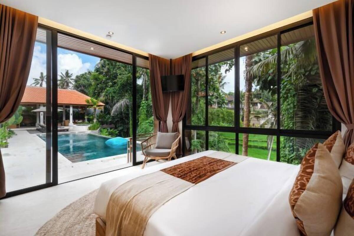 A bedroom with a large bed, a chair, a TV, and a view of a swimming pool and greenery.